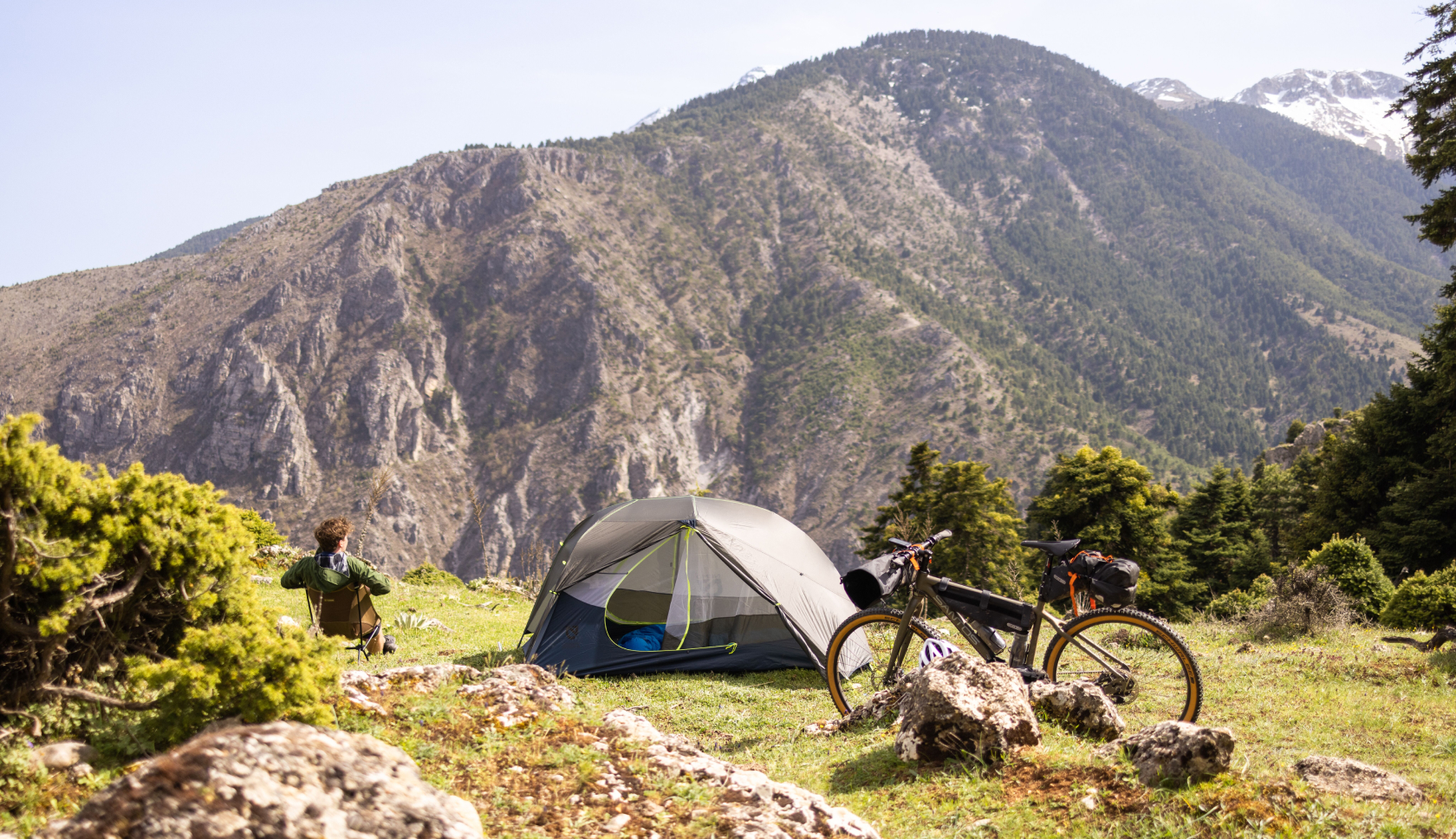 Een persoon zit tijdens bikepacking op een campingstoeltje in de bergen naast zijn tentje en fiets.