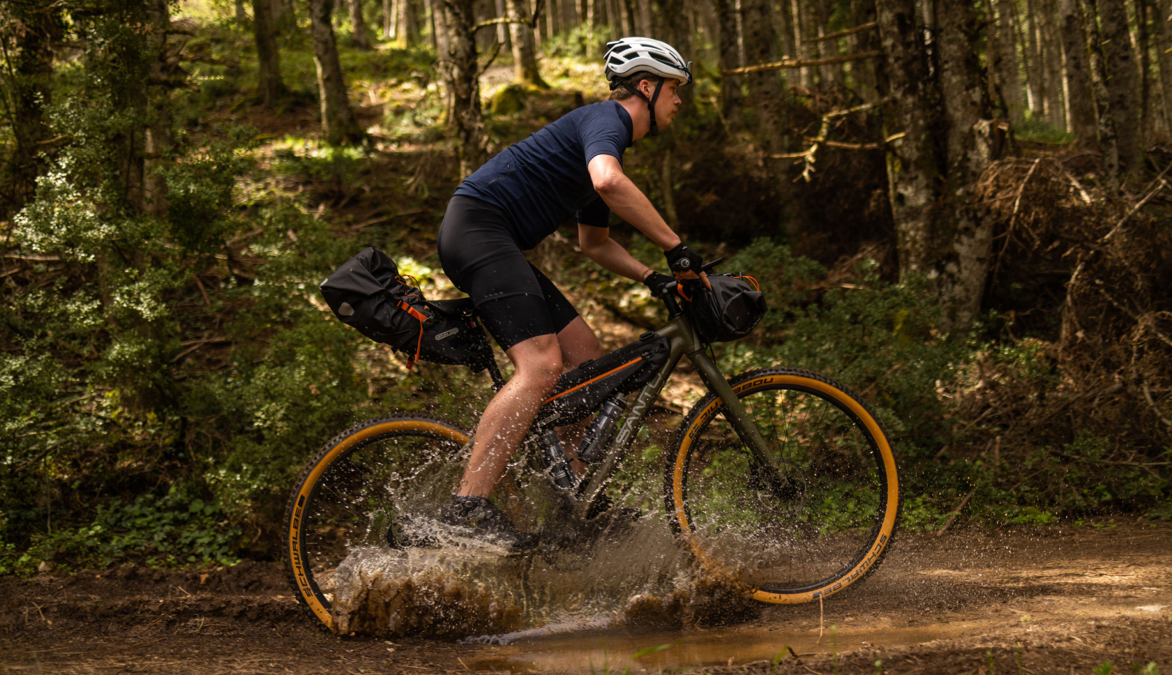 Een bikepacker fietst door een plas in het bos en water spat op.